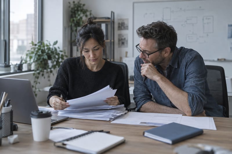 Two colleagues working through a stack of paper forms beside a laptop